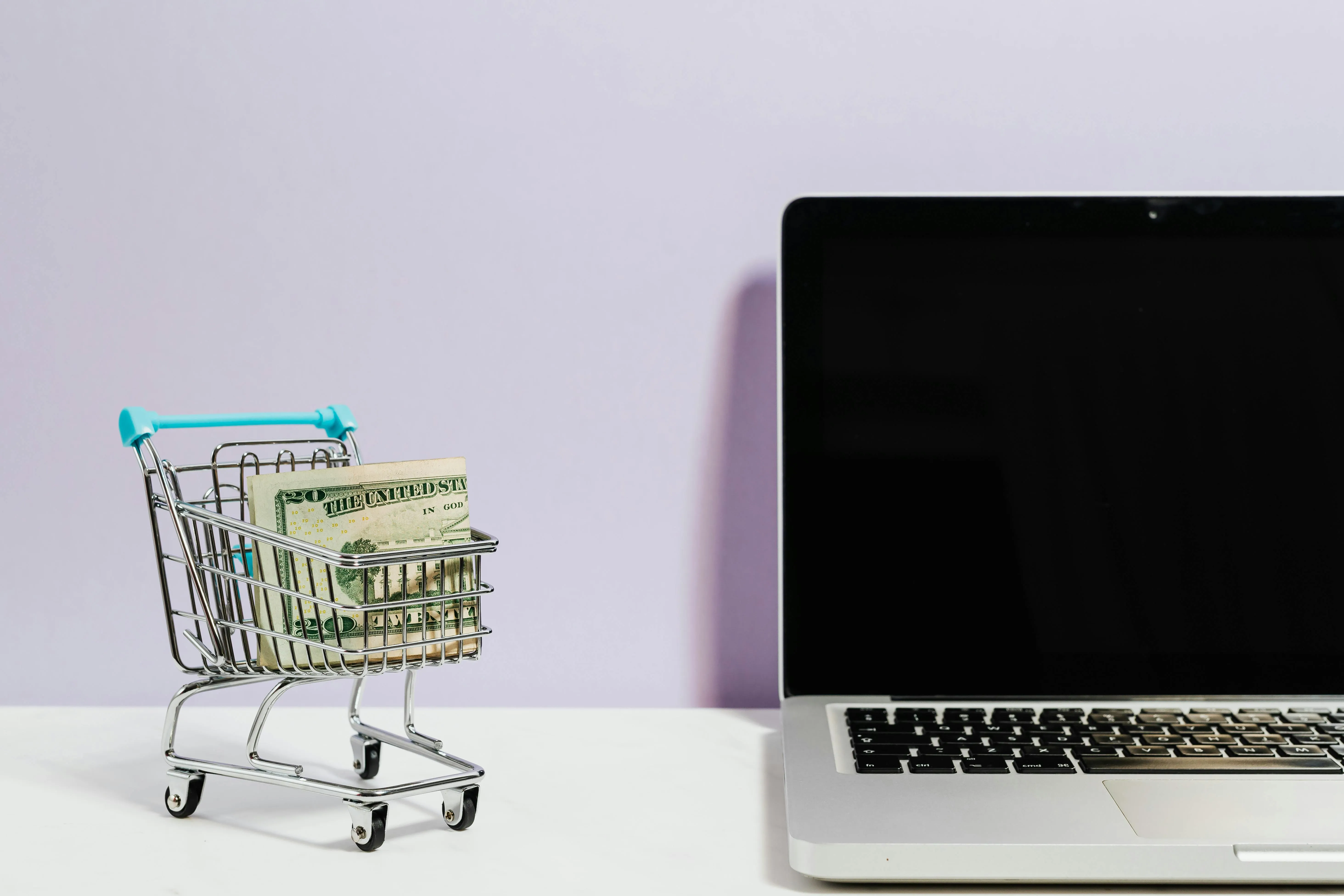 Small shopping cart carrying dollar bills next to a laptop on pink background representing online e-commerce revenue